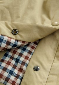 Giacca di tessuto beige con texture in corden di color sabbia chiaro, con fodera interna a quadri nei colori rosso, blu e crema, e bottoni a pressione in metallo.