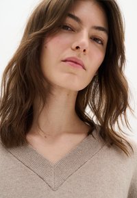 Beżowy sweter z dekoltem w serek o ryflowanej strukturze. Miękka tkanina, luźny krój. Włosy ciemnobrązowe, falowane, z naturalną objętością i subtelnym połyskiem.
