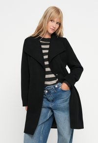 Cappotto di lana nero con un ampio colletto e apertura frontale, abbinato a un maglione a strisce grigie e nere, e jeans a vita alta di colore azzurro chiaro.