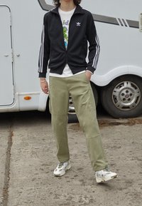 Person som bär en svart Adidas träningsjacka, en grafisk vit T-shirt, ljusgröna byxor och vita sneakers, stående på betong nära ett vitt fordon.