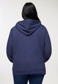 Donna con lunghi capelli neri, che indossa un semplice hoodie blu navy e jeans blu scuro, di spalle contro uno sfondo chiaro.