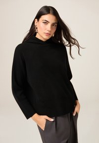 Czarna bluza z kapturem, zakończona prążkowanym dołem i mankietami, zestawiona z szarymi spodniami. Tkanina wygląda na miękką, a krój jest luźny.