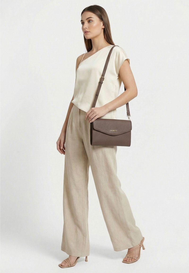 Mujer con top crema de un solo hombro y pantalones beige de pierna ancha, llevando un bolso marrón de hombro y sandalias beige con tacón, posando sobre un fondo blanco.