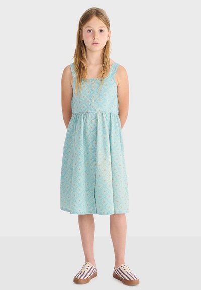 Dievča s dlhými vlasmi v bezrukávových svetlomodrých šatách s potlačou a pruhovaných teniskách, stojaca s rukami za chrbtom.