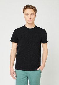 Schwarzes T-Shirt mit kurzen  Ärmeln in strukturierter Optik und rundem Ausschnitt, kombiniert mit hellgrünen Hosen. Detail mit Fronttasche auf der linken Seite.