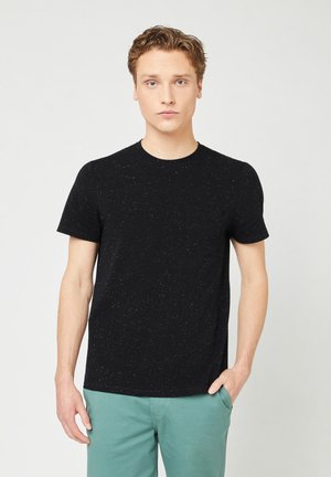 T-shirt nera a maniche corte con texture maculata e scollo rotondo, abbinata a pantaloni verde chiaro. Dettaglio con tasca frontale sul lato sinistro.