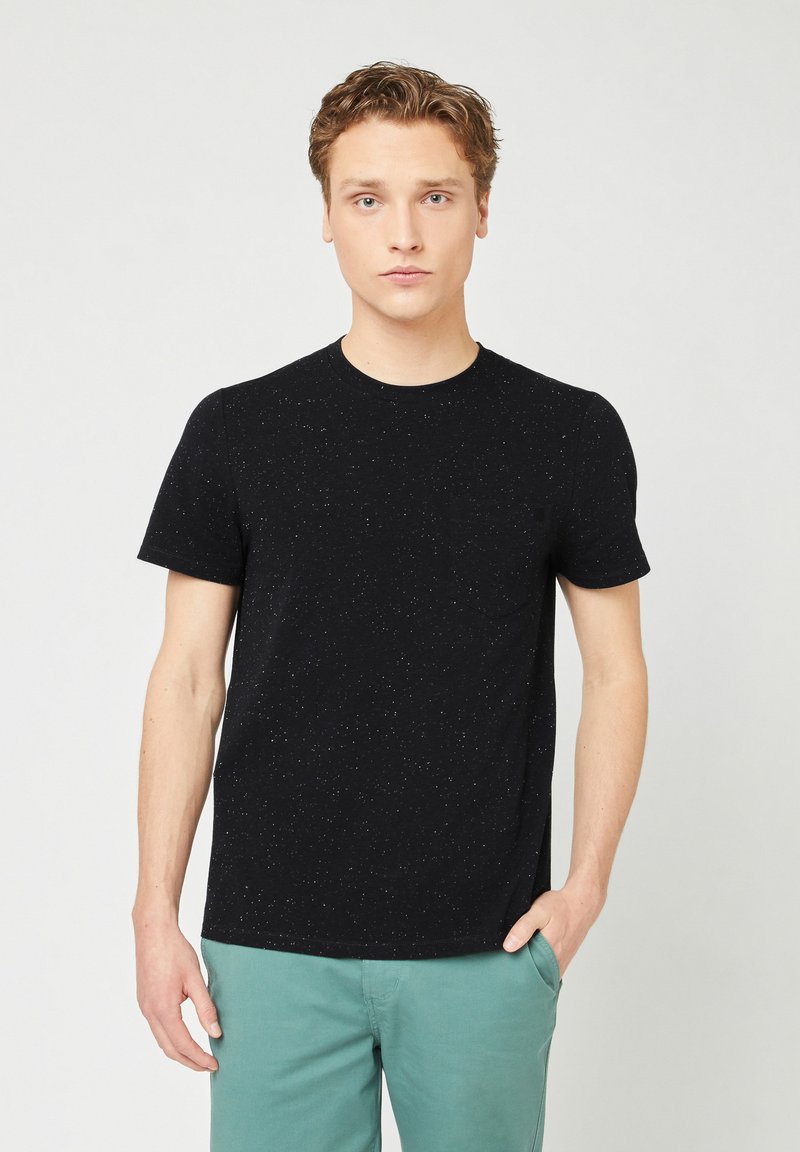 Schwarzes T-Shirt mit kurzen Ärmeln in strukturierter Optik und rundem Ausschnitt, kombiniert mit hellgrünen Hosen. Detail mit Fronttasche auf der linken Seite.