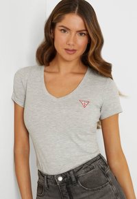 Light grey V-neck t-shirt in soft fabric with short sleeves, featuring a red Guess logo patch on the left chest. Worn with dark high-waisted jeans.