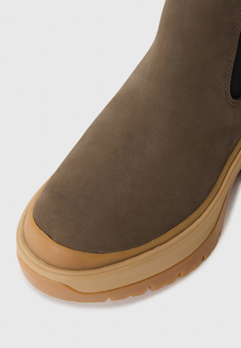 Botín de ante marrón con suela de goma. Punta redondeada, textura suave y paneles laterales elásticos. Detalles cosidos a lo largo de los bordes.