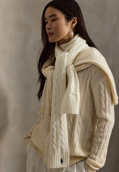 Kremfarget kabelstrikket genser med ribbestrikkede mansjetter, lagdelt med et matchende skjerf. Inkluderer en liten brodert logo nær kanten. Elegant, myk tekstur.