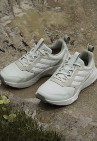 Ζευγάρι ανοιχτόχρωμων γκρίζων παπουτσιών Adidas Terrex για ορεινό τρέξιμο πάνω σε λασπώδες έδαφος κοντά σε χορτάρι και μια μικρή λακκούβα.
