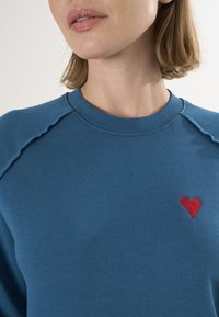 Niebieska bluza z okrągłym dekoltem, z haftem czerwonego serca na piersi i teksturowanym, lekko postrzępionym szwem na ramieniu.