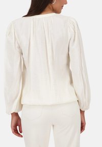 Beige-bluse aus leichtem stoff, mit langen puffärmeln und gerafftem rückendetail. Getragen mit hellen hosen.