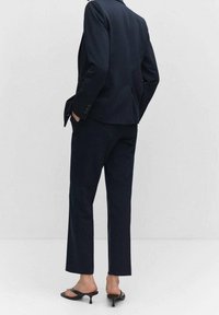 Costume bleu marine sur mesure avec pantalon ajusté et blazer ; tissu lisse ; léger éclat ; fermeture à un bouton ; ballerines pointues.