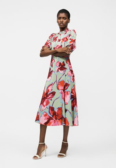 Γυναίκα που στέκεται με σταυρωμένα τα χέρια, φορώντας ένα πράσινο φόρεμα μέσης με μεγάλα κόκκινα και ροζ λουλουδένια μοτίβα και λευκά πέδιλα με ψηλά τακούνια.