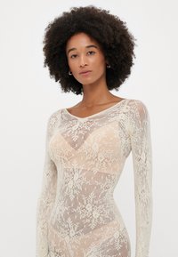 Δαντελένιο κορμάκι με V-λαιμόκοψη, μακριά μανίκια, σε κρεμ απόχρωση με λουλουδάτα σχέδια και ημι-διάφαστη υφή.