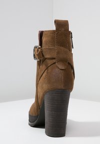 Brune ruskind ankelstøvle med høj stablet hæl, ankelrem og spænde detalje. Tekstureret bag og glat mørk ydersål.