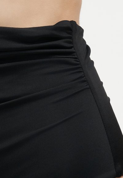 Κοντινή λήψη μαύρου ραφτά υφάσματος που δείχνει λεπτομέρεια πλαϊνής ραφής σε εφαρμοστά ρούχα πάνω σε ανοιχτόχρωμο δέρμα και λευκό φόντο.