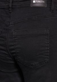 Pantalones de mezclilla negra con un diseño de bolsillo trasero. La textura es suave con costuras visibles a lo largo de las costuras y bordes. La etiqueta de la marca está attached.
