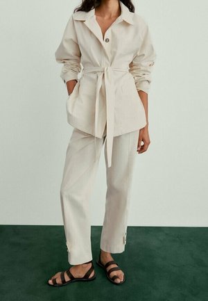 Beżowa kurtka z paskiem i guzikiem, zestawiona z pasującymi spodniami. Teksturowana tkanina, otwarty kołnierz oraz czarne sandały na paski dopełniają strój.