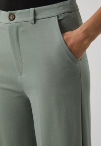 Spodnie w jasnozielonym materiale o gładkiej teksturze, z zapięciem na guzik, przednimi kieszeniami i dopasowanym krojem.