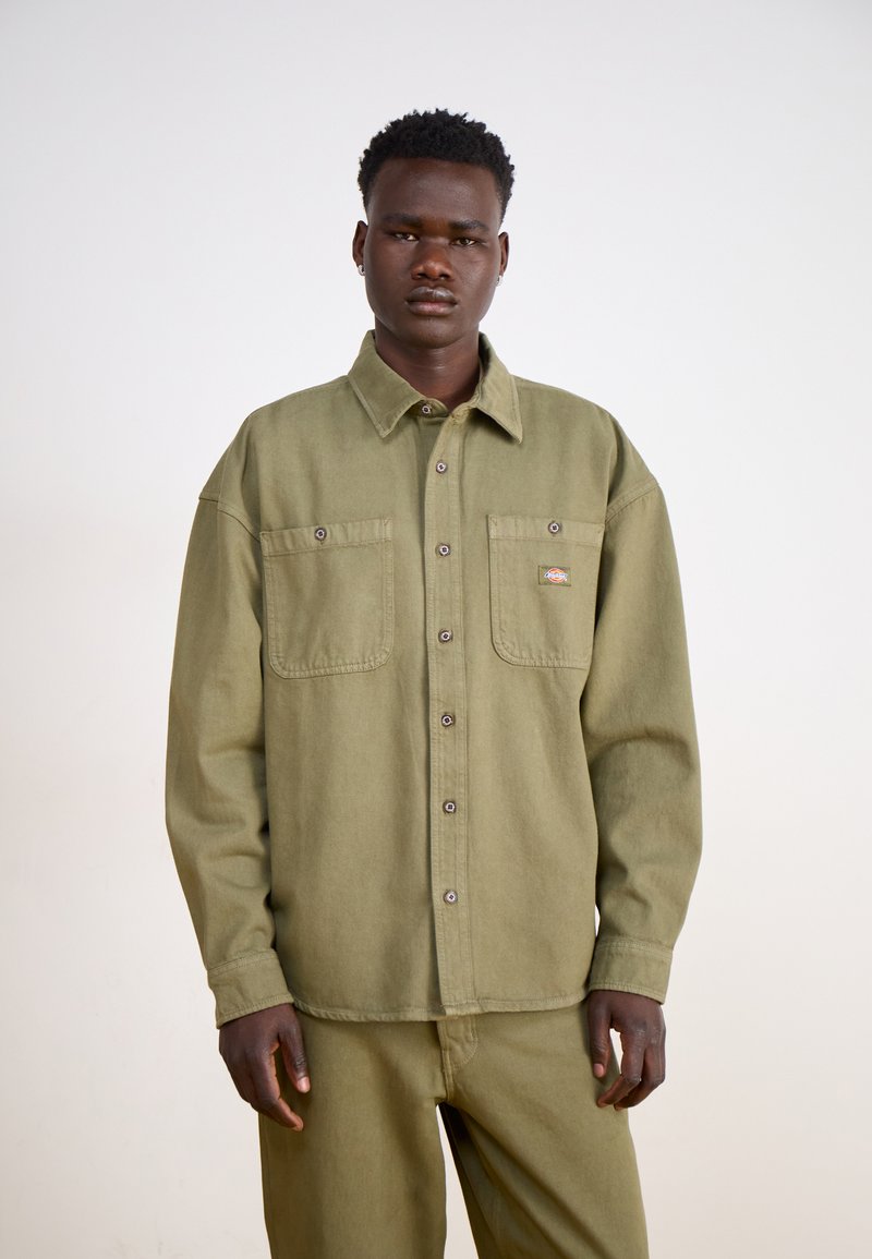 Camisa de algodón verde oliva con botones, que presenta dos bolsillos en el pecho, un cuello de punta y botones de metal, usada con pantalones a juego.