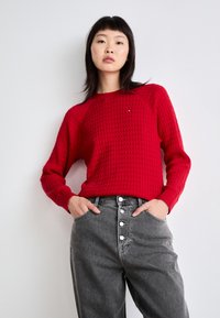 Suéter rojo de punto con un patrón texturizado, cuello redondo y puños acanalados, combinado con pantalones de mezclilla grises de tiro alto con cierre de botones.