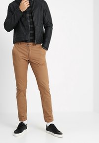 Musta vetoketjullinen nahkatakki mustan ja harmaan ruudullisen paidan päällä, yhdistetty tanin värisiin slim fit -chinoihin ja mustiin tennareihin.