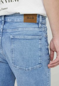 Jean en denim bleu clair avec étiquette en cuir marron BZB à la taille. Présente une poche arrière avec des bords cousus et une texture lisse.