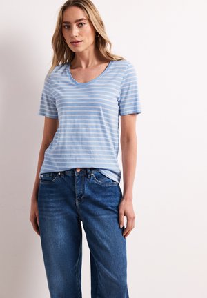 Jeune femme portant un T-shirt rayé bleu clair et blanc et un jean bleu, debout devant un fond clair uni.