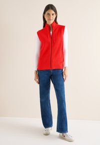 Rotes, geripptes Reißverschlussweste mit hohem Kragen, kombiniert mit hellblauen Schlagjeans und weißen Turnschuhen. Heller Hintergrund.