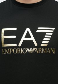 Fekete ing nagy, fehér és arany színű "EA7 Emporio Armani" felirattal és arany sas logóval a mellkas közepén.