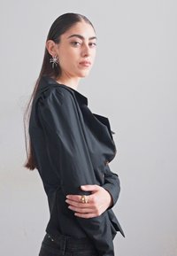 Svart strukturerad blus med puffärmar, prydd med bruna knappar, bärs med mörka jeans. Modellen visar en profilbild, minimalistisk bakgrund.