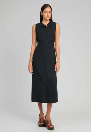 Femme portant une robe midi noire sans manches avec un nœud au niveau de l'encolure et des talons compensés marron à bout ouvert, debout devant un fond uni.