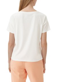 Weißes T-Shirt mit kurzen Ärmeln und rundem Ausschnitt aus glattem Material. Getragen mit pfirsichfarbenen Hosen, die Rückansicht wird gezeigt.