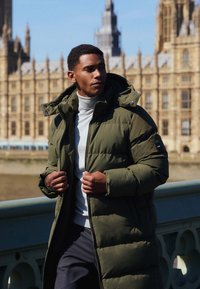 Olivově zelená péřová bunda s kapucí, zipem a bočními kapsami, nošená přes světle šedý rolák, na pozadí historického prostředí.