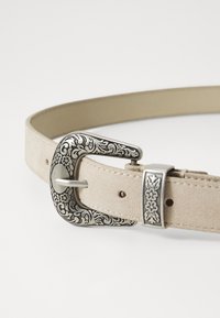 Ljust beige mockaskärp med en utsmyckad silverspänne som har blommotiv och en metallisk detalj. Slät yta och justerbar design.