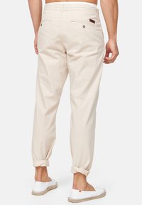Man in beige opgerolde broek en witte instapschoenen, staande met de rug naar de camera op een effen lichte achtergrond.
