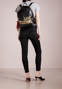 Žena v černých vysokých podpatcích mules a úzkých černých džínách s šedým tričkem, nese černý batoh "LOVE MOSCHINO".