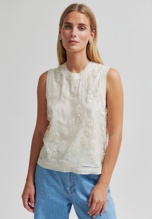 Ung kvinde iført hvid ærmeløs top med blomsterformede palietdetaljer og lyseblå jeans, stående foran en ensfarvet baggrund.