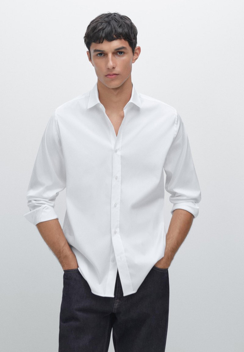 Biała koszula z długim rękawem i zapięciem na guziki, z kołnierzykiem typu spread, dopasowany krój i podwinięte rękawy, zestawiona z ciemnymi dżinsami. Minimalistyczny design.