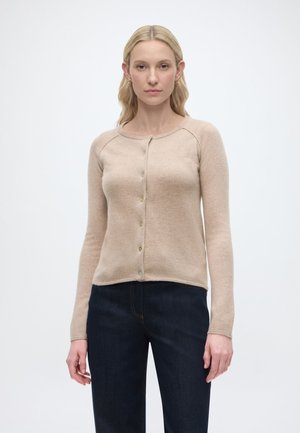 Frau mit blondem Haar, die eine beige, geknöpfte Strickjacke und dunkelblaue Jeans trägt, steht vor einem einfachen hellen Hintergrund.