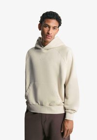 Béžová mikina s kapucí, s klokankou, žebrovanými manžetami a lemem, s decentním vyšívaným logem na prsou.