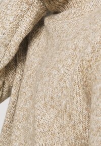 Pull beige en maille avec une finition texturée, présentant une coupe ample et des poignets côtelés proéminents. Le matériau semble doux et chaud.