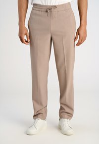 Mężczyzna w beżowych spodniach na sznurek i białych trampkach stoi na jednolitym, jasnym tle.