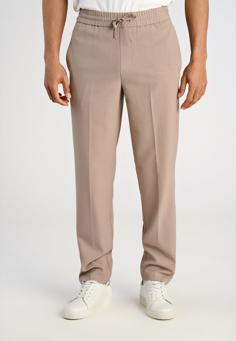 Mężczyzna w beżowych spodniach na sznurek i białych trampkach stoi na jednolitym, jasnym tle.