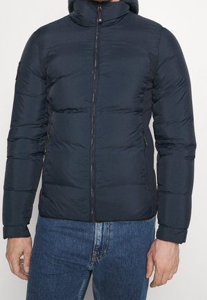 Mand iført en navyblå polstret jakke med frontlynlås og lommer med lynlås i siderne, kombineret med blå jeans.