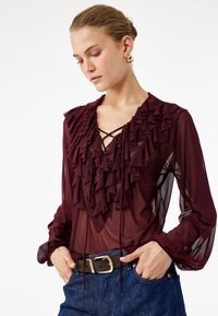 Donna che indossa una blusa trasparente bordeaux con volant, scollo all'americana in pizzo e maniche a sbuffo, abbinata a jeans blu e una cintura marrone.