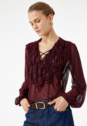 Donna che indossa una blusa trasparente bordeaux con volant, scollo all'americana in pizzo e maniche a sbuffo, abbinata a jeans blu e una cintura marrone.