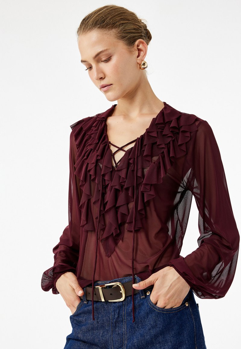 Donna che indossa una blusa trasparente bordeaux con volant, scollo all'americana in pizzo e maniche a sbuffo, abbinata a jeans blu e una cintura marrone.